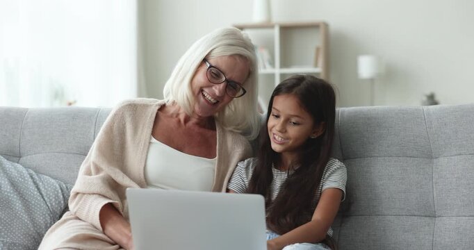 Cheerful blonde grandmother and preteen little schoolkid girl reading text aloud on laptop screen, chorusing poem, song lyrics, beating out rhythm with hands, speaking, singing, smiling, laughing