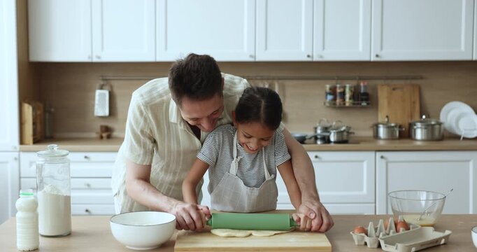 Happy baker dad and adorable daughter kid in apron baking together, rolling dough in home kitchen table, touching faces of each other with floury fingers, having fun, laughing