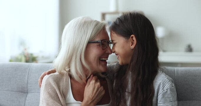Cheerful blonde grandmother and cute loving granddaughter kid hugging with noses touch, enjoying closeness, family relations, affection, sitting on home couch, smiling, laughing