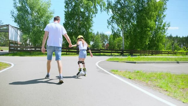 Caring father securing happy daughter, holding her hands helping to maintain balance on roller skates while teaching child rollerblading in summer park.