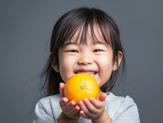 A young Asian girl smiles brightly as she holds a ripe orange in her hands.