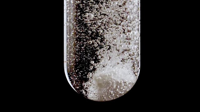 Close up tablet of aspirin, vitamin falls into of water in glass test tube in slow motion on black background. Macro shot of bubbles effervescent. The medicine concept, health care, treatment.FullHD 