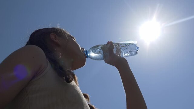 The girl drinks clean, cool water from a bottle. Drink water in the heat against the background of the sun and sky.