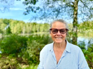 A portrait of a older smiling woman wearing glasses, outside in the Swedish nature