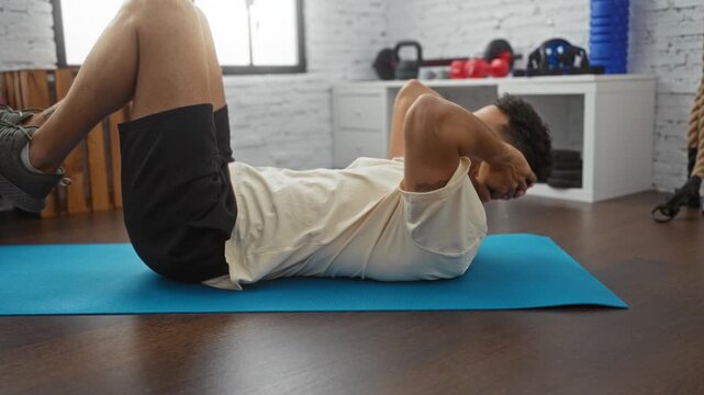 A young hispanic man performing sit-ups on a blue mat in an indoor gym setting, emphasizing fitness and well-being.