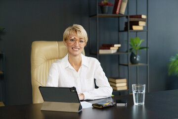 A smiling woman dressed in a white shirt engages with her tablet at a stylish office desk, surrounded by books and a refreshing glass of water.