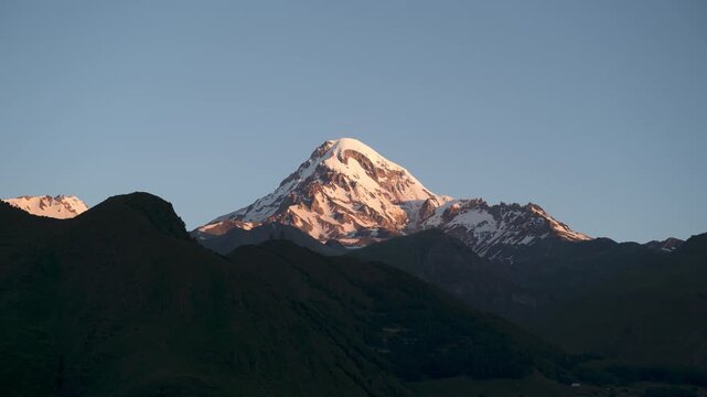 Timelapse shows Kazbegi peak and Gergeti Trinity Church with starry sky and moonrise. Mountains glow at sunrise. Scenic Georgia landscape in night-to-day transition.