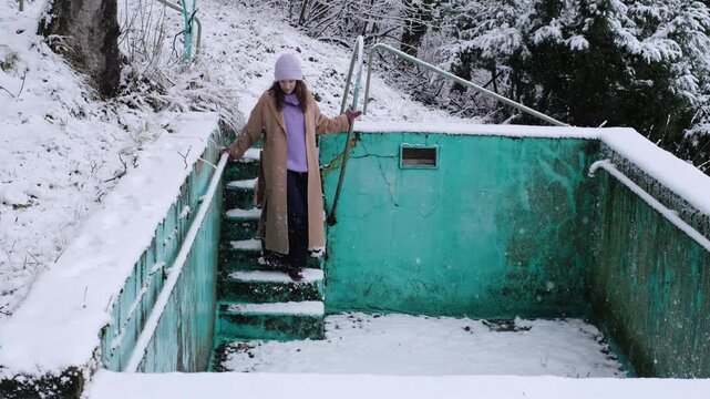 Attractive woman pushing off the snow from a railing in an abandoned swimming pool. Insecure what to do, everyday problems. White Christmas season, adult playing with falling snow in winter wonderland
