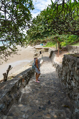 A lady in a large hat stands in the shade of a tropical tree on the stone steps of a staircase leading up a hill and takes photos on her phone.