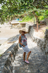 A lady in a large hat and glasses stands in the shade of a tropical tree on the stone steps of the stairs leading up the hill.