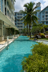 View of a blue water pool in the courtyard of a tropical resort. Lush tropical vegetation grows around the pool. At the edge of the pool there are metal stairs to get into the water.