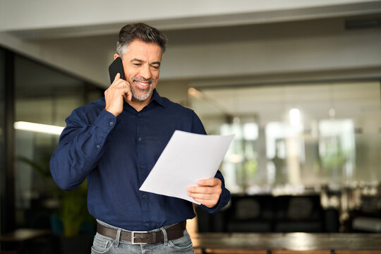 Happy busy mature older business man holding papers making call at work. Smiling middle aged senior professional businessman executive looking at documents talking on cell phone standing in office.
