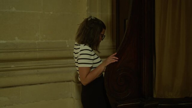 Young hispanic woman in striped shirt standing in a church in italy, near a large wooden confessional booth, immersed in the serene and historical christian setting of the cathedral.