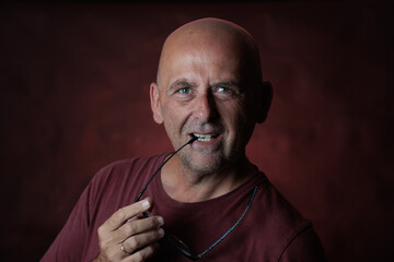 Portrait of a smiling mature man holding his glasses with teeth on red background