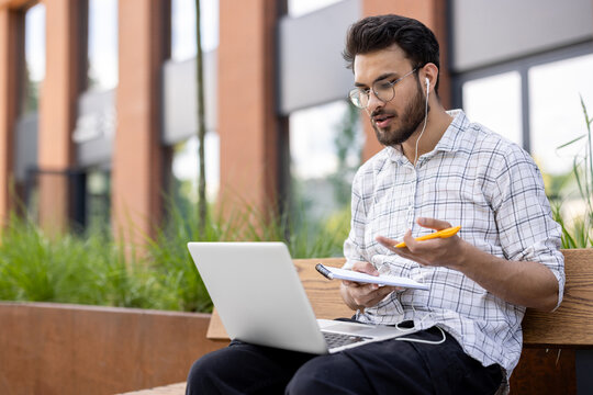 Young man taking notes during an online class on laptop outdoors