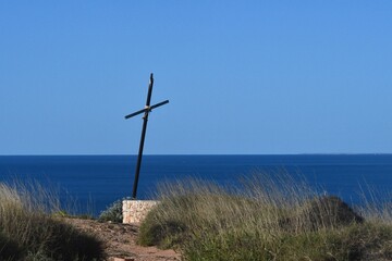 A wooden cross blackened with age and tied together with rope stands on a deserted headland. It rests on sandstone, and is surrounded by wild grasses. It is seen against a blue sea and sky.