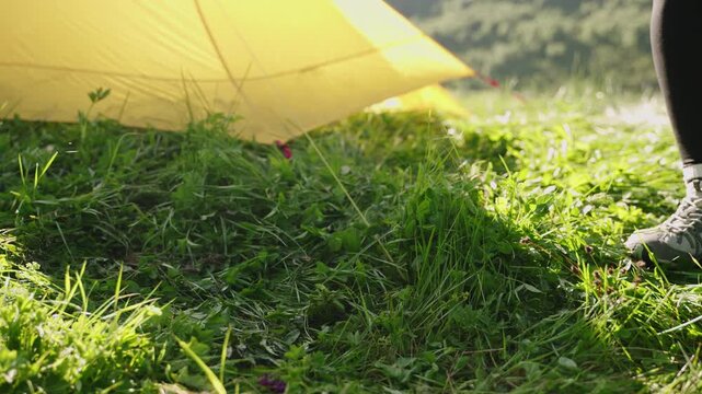 Woman places portable mini gas stove, sets cooking pot near yellow tent. Slow motion scene of backpacking trip showing ultralight gear in. Outdoor adventure in nature setting, preparing to boil water.