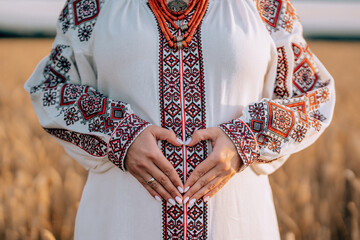 Pregnant woman showing heart-shaped symbol gesture on belly, wheat natural field