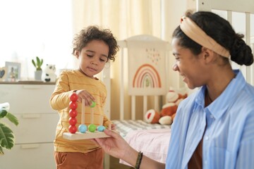 Smiling woman engaging young child with colorful educational toy featuring wooden beads in cozy, well-decorated nursery adorned with pastel rainbow accents