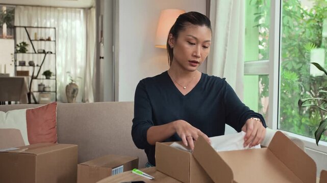 Medium shot of young Asian woman opening cardboard box with new pair of leather shoes being delivered to her home