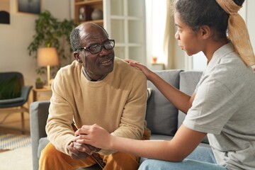 Elderly man sitting in living room being comforted by young African American girl holding his hand...