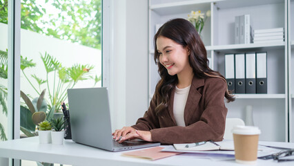 A young businesswoman, dressed in a brown jacket and beige sweater, is smiling while working on her laptop in a bright, modern home office.