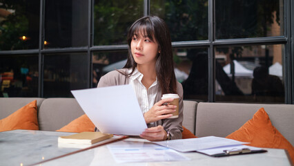 Businesswoman is focused on reviewing documents while seated in a modern, stylish cafe.