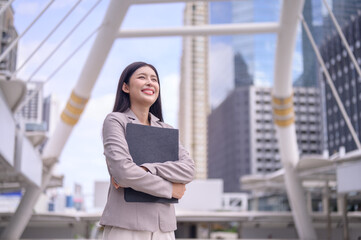 young professional woman standing outdoors in a modern urban environment, smiling confidently as she holds a black folder close to her chest.