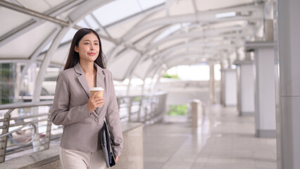 Young woman walking through a modern, covered walkway or pedestrian bridge, holding a cup of coffee in one hand and a tablet or folder in the other.