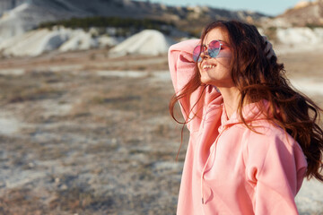 stylish woman in pink hoodie and sunglasses poses confidently in desert landscape with mountains