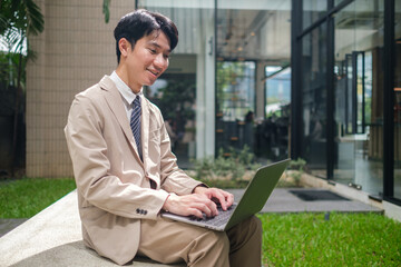 Smiling young businessman sitting comfortably in a modern outdoor office, working on his laptop.