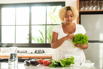 Young African American curvy woman standing in the kitchen preparing a salad of green lettuce leaves, different vegetables on the table. Diet and healthy lifestyle. Body positive.