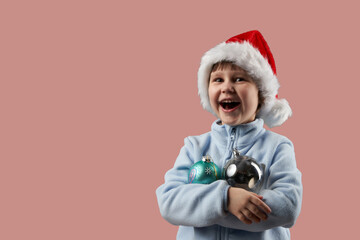 Laughing child in Santa Claus hat clutching glass balls to his chest to decorate Christmas tree. The background is pink. Copy space