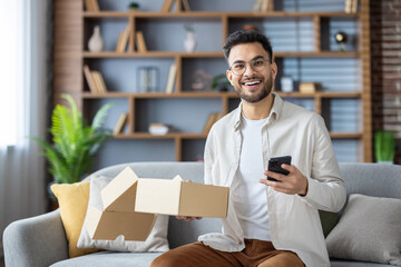 Portrait of a young Muslim man in glasses sitting happy at home on the couch, holding a phone and an open parcel box, looking and smiling at the camera