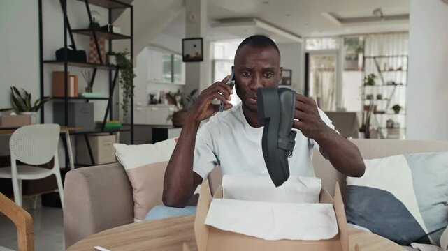 Medium shot of black man calling to customer service on smartphone when getting wrong pair of shoes in parcel