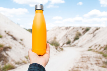 Male hand holding steel thermo water bottle of yellow color on background of sand hills.