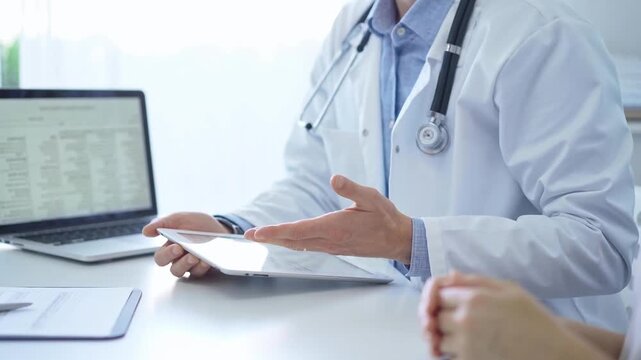 Doctor and a patient. The physician, wearing a white medical coat over a blue shirt, is using tablet computer during a consultation in the clinic. Medicine and science