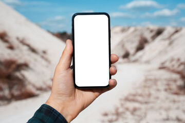 Close-up of male hand holding smartphone with blank on screen, mockup, on background of sand hills.