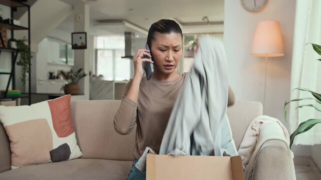 Medium shot of female Asian customer calling online store while receiving defective clothes in her package delivered to home