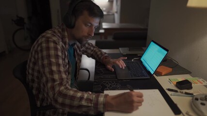 Focused man in headphones with a laptop late at night at home, taking notes, student learning a language, watching an online webinar, hearing an audiocourse, the concept of e-education. Homework. 