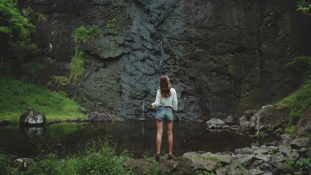 Young woman stands near a waterfall pool in a lush jungle, touching her long hair, enjoying the serene setting of french polynesia's moorea island