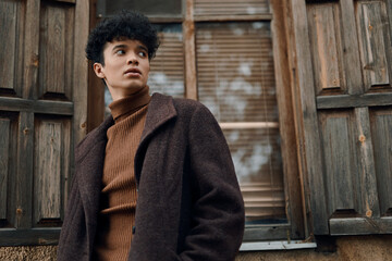 Fototapeta premium Fashionable young african american woman in a brown coat posing in front of an old building with wooden shutters and a door