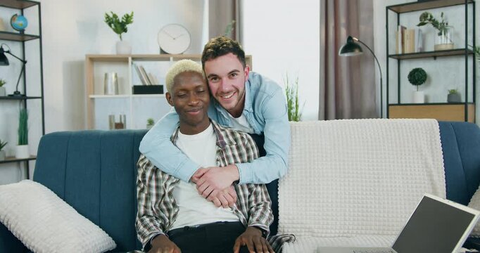 Young men standing behind the sofa and hugging his african american partner which sitting on the sofa and they both looking into camera in cozy living-room