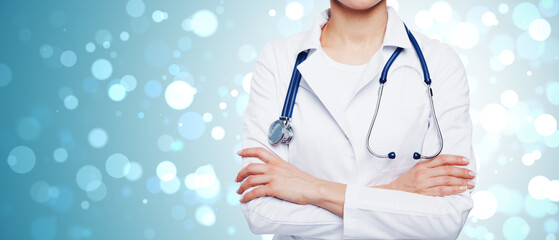 Portrait of female doctor wearing white coat and stethoscope with arms crossed on blue bokeh...