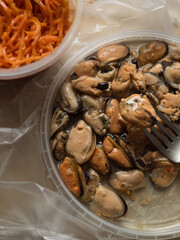 Vertical Flat Lay of Two Plastic Containers with Korean Carrot Salad and Marinated Mussel Flesh on Polyethylene Surface.