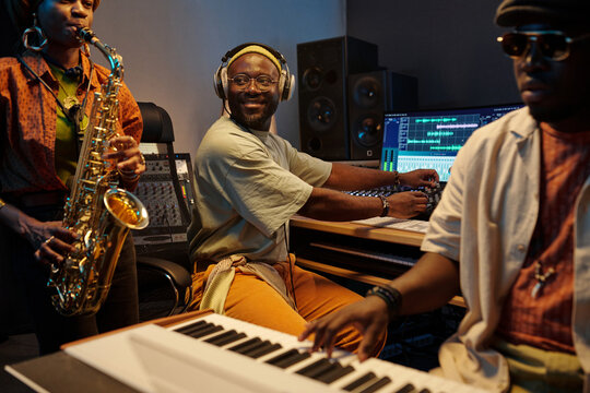 Musicians performing in professional recording studio environment with various audio equipment surrounding recording engineer focused on mixing tracks