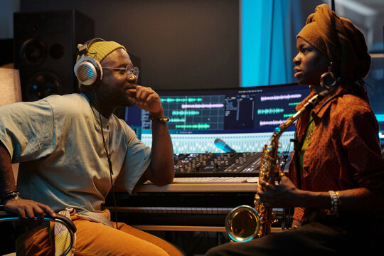 Two musicians wearing headphones working in recording studio with digital software while discussing and playing instruments including saxophone and keyboard with electronic mixers surrounding