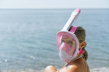 FA woman wearing a pink snorkel mask is sitting on a beach. The water is calm and the sky is clear.