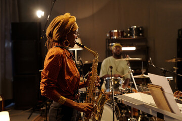 Musicians rehearsing jazz performance with saxophonist in foreground and drummer in background. Recording studio setting with instruments and equipment surrounding musicians - Powered by Adobe