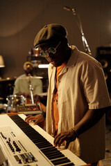 African American musician playing keyboard in music studio setting while wearing sunglasses and hat with another band member in background on-stage performing